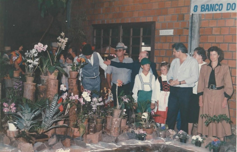 Outro instante da visitação pública a Exposição. 
Da direita à esquerda:  casal Elizabete e Werner Wanderer, tendo ao fundo a senhora Reinilda (Frühauf) Fuck;  Reinoldo Fuck, tendo a frente dois de seus netos) admirando um Cymbidium híbrido e conversando com vice-prefeito Verno Scherer; e o casal Dieter Leonard e Vera Seyboth admirando um exemplar de híbridos de diversos gêneros. 
Imagem: Acervo Memória Rondonense. 