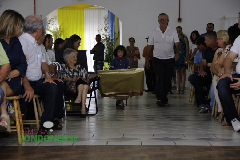 João Pedro, filho do prefeito empossado Márcio Andrei Rauber trazendo a peça artística produzida pelo agricultor e artista Jão Zimmermann, de caminha branca, caminhando ao lado, à direita. 
A senhora sentada, à esquerda, em cadeira dobrável, é Amabile Brambati, avó materna do prefeito Márcio. 
Imagem: Acervo Memória Rondonense - Crédito: Tioni de Oliveira 