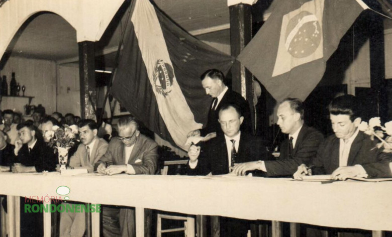 Componentes da Mesa de Honra assinando o termo e ata de posse. Ao tempo em que o senhor Arlindo Alberto Lamb repassa o seu discurso de posse. 
Da esquerda à direita, sentados: 1º - pastor Joachim Pawelke (de terno escuro); 2º - Wilson Ballão, juiz de direito da comarca de Toledo; 3º - não identificado (de óculos); 4º - vereador Helmuth Priesnitz, presidente da Câmara Municipal de Marechal Cândido Rondon; 5º - Wilson Carlos Kuhn, da Prefeitura Municipal de Toledo; e 6º - não identificados. 
Imagem: Acervo idem 
